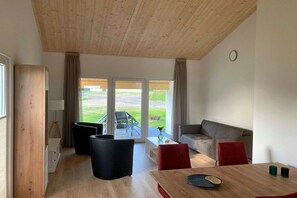Ein modernes Wohnzimmer mit Holzboden, einer Couch, einem roten Sessel, einem Esstisch und Blick ins Freie durch große Glaswände.