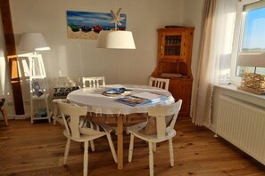 A dining area with a round table, white chairs, a wooden cabinet, and a painting of boats on the wall. - North Sea house with pirate's nest (Nordstrand)