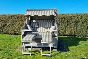 Un espace extérieur abrité, avec un banc rembourré et une table, le tout entouré d’une haie.