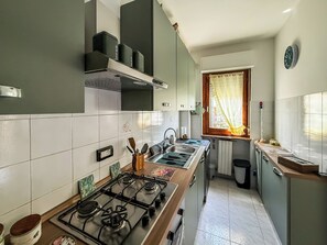 A compact kitchen with a gas stove, a sink, and a window with curtains. - Casa Armando (Passignano sul Trasimeno)