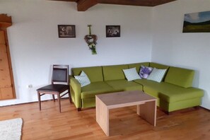 Ein Zimmer mit einem grünen Sofa, einem Holztisch und einem Stuhl. An der Wand hängen zwei gerahmte Bilder und ein Wandkranz.