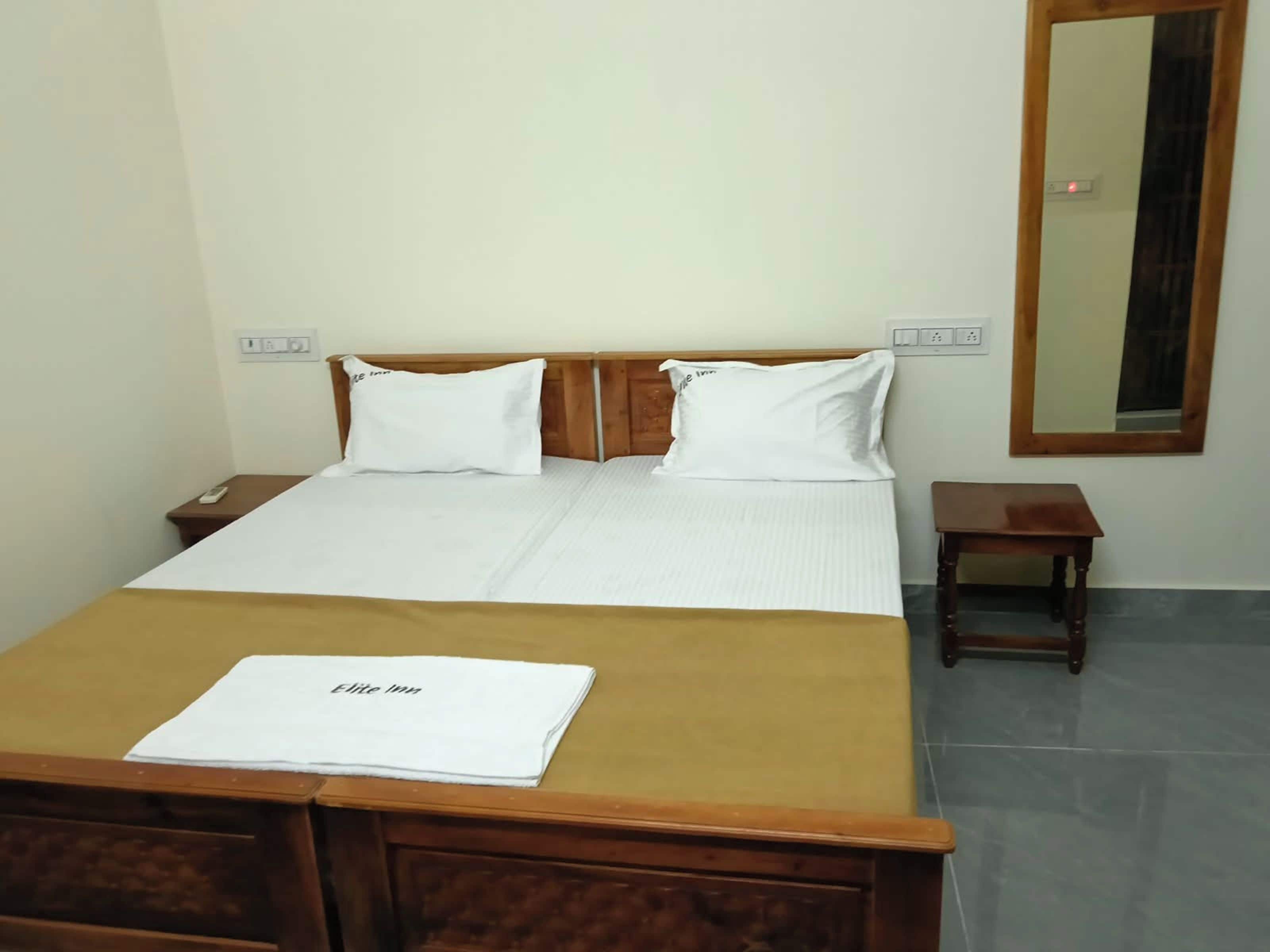 A hotel room with a bed, a bedside table, a nightstand, a wooden headboard, a mirror, and a sign indicating 'Elite Inn'.