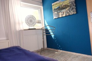 A bedroom with a bed, a radiator, a window with curtains, a wall-mounted circular light, and a poster. - Holiday apartment with 2 bedrooms (Kappeln-Lüttfeld)