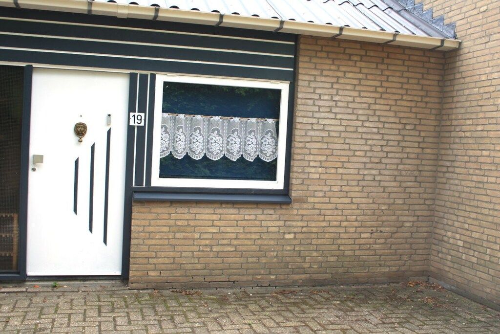 A building with a white door marked with the number 19, a window with lace curtains, and a brick wall.