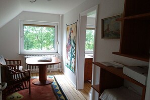 Um quarto com uma mesa de madeira, cadeiras, um radiador, uma janela com vista para árvores, uma tapeçaria colorida e um quadro na parede.