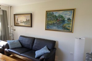 A living room with a black leather sofa, two blue cushions, a lamp, and two framed paintings on the wall. - Beach holiday at the "Kiebitz" (Bensersiel)