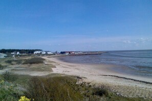 Eine Küstenlandschaft mit Sandstrand, einer hölzernen Pier, die ins Wasser führt, und einer Reihe Häuser im Hintergrund.