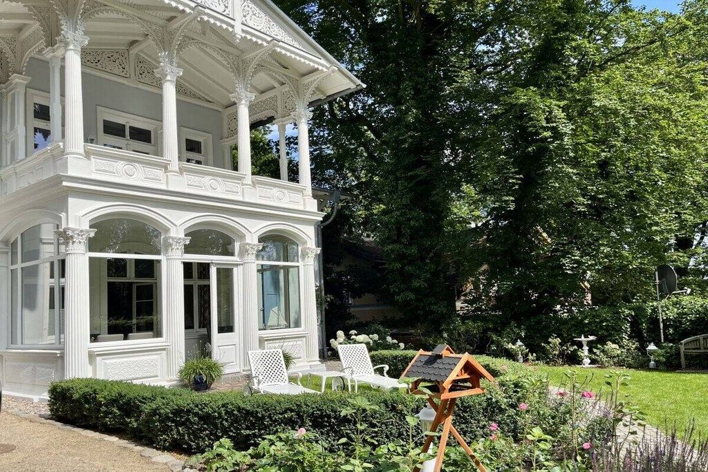 A white gazebo with ornate columns and a glass enclosure, surrounded by a well-maintained garden with various plants and a wooden easel.