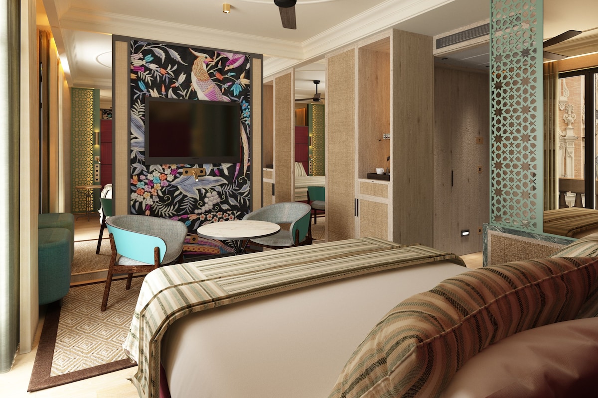 Een hotelkamer met een bed, twee fauteuils, een klein rond tafeltje, een flatscreen televisie bevestigd aan een muur met bloemenmotief en een plafondventilator.