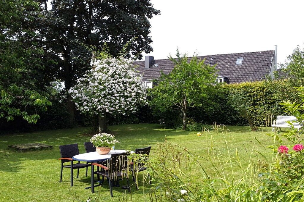 A garden with a table and chairs, surrounded by greenery and a house in the background.