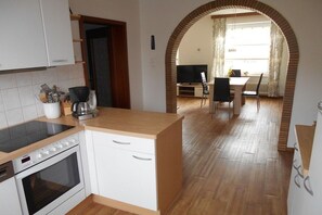 A kitchen with a stove, oven, and white cabinets. There is a dining area with a table and chairs, and a TV in the background. - Holiday apartment with 2 bedrooms (Flensburg)