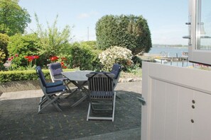 An outdoor seating area with a table and chairs, surrounded by lush greenery and flowers, overlooking a body of water. - Holiday apartment with 1 bedroom (Stexwig)