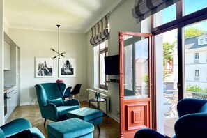 A modern living room with a teal armchair, ottoman, and a television on a stand. There are framed pictures on the wall, a small table with a vase, and a large window with a view of buildings outside. - Villa Kramme - close to the beach & central (Seebad Heringsdorf)