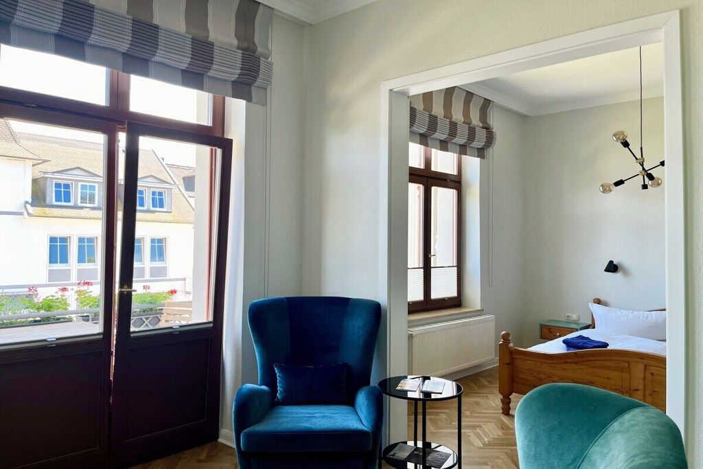 Habitación con cama, una silla, una mesita y ventana con vista a edificios.