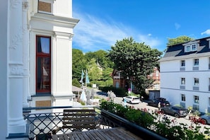 A balcony with a wooden bench, overlooking a street with parked cars and a building with a red door. - Villa Kramme - close to the beach & central (Seebad Heringsdorf)