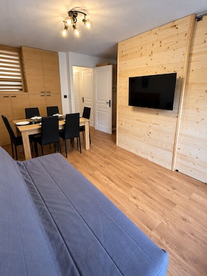 A room with a dining table, chairs, a sofa, a TV mounted on a wooden wall, and a ceiling light fixture. - Apartment with a balcony and a heated swimming pool (Allos)