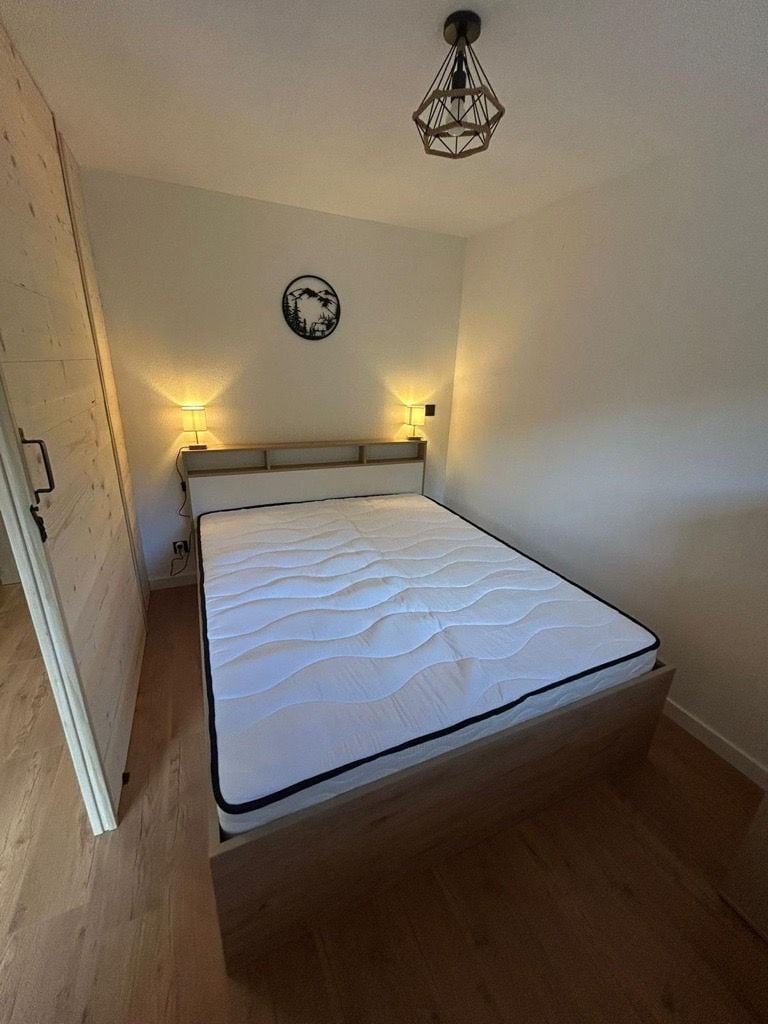 A single bed with a wooden frame and a white mattress in a room with wooden flooring and white walls.