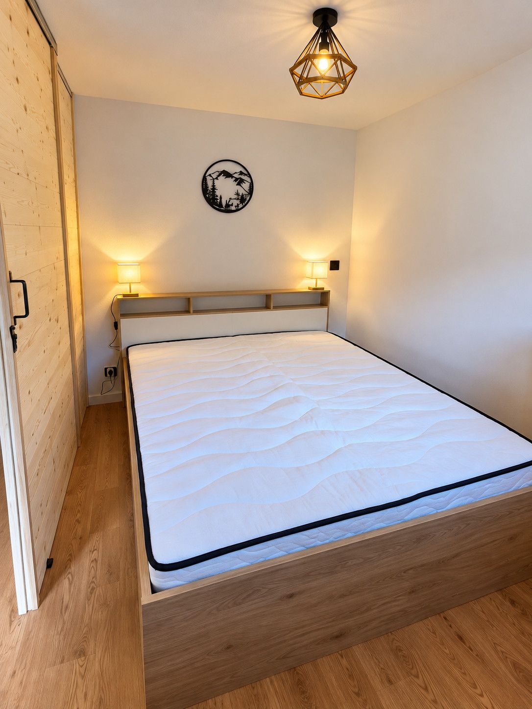 Una cama individual con estructura y cabecero de madera, dos lámparas de noche, un aplique de pared y un adorno mural.
