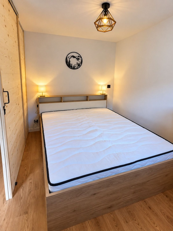 Uma cama de solteiro com estrutura e cabeceira de madeira, duas luminárias de apoio, um ponto de luz fixado na parede e um adorno de parede.