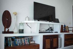 Een woonkamer met een flatscreen televisie bevestigd aan een witte plank, een houten klok, een vaas met bloemen en een boekenkast vol boeken.