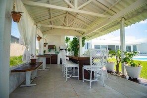 A covered patio area with a dining table and chairs, potted plants, and a view of a swimming pool.