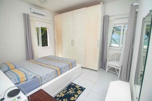 A bedroom with a bed, a desk with a computer, a chair, a wardrobe, and a window with a view of the sea.