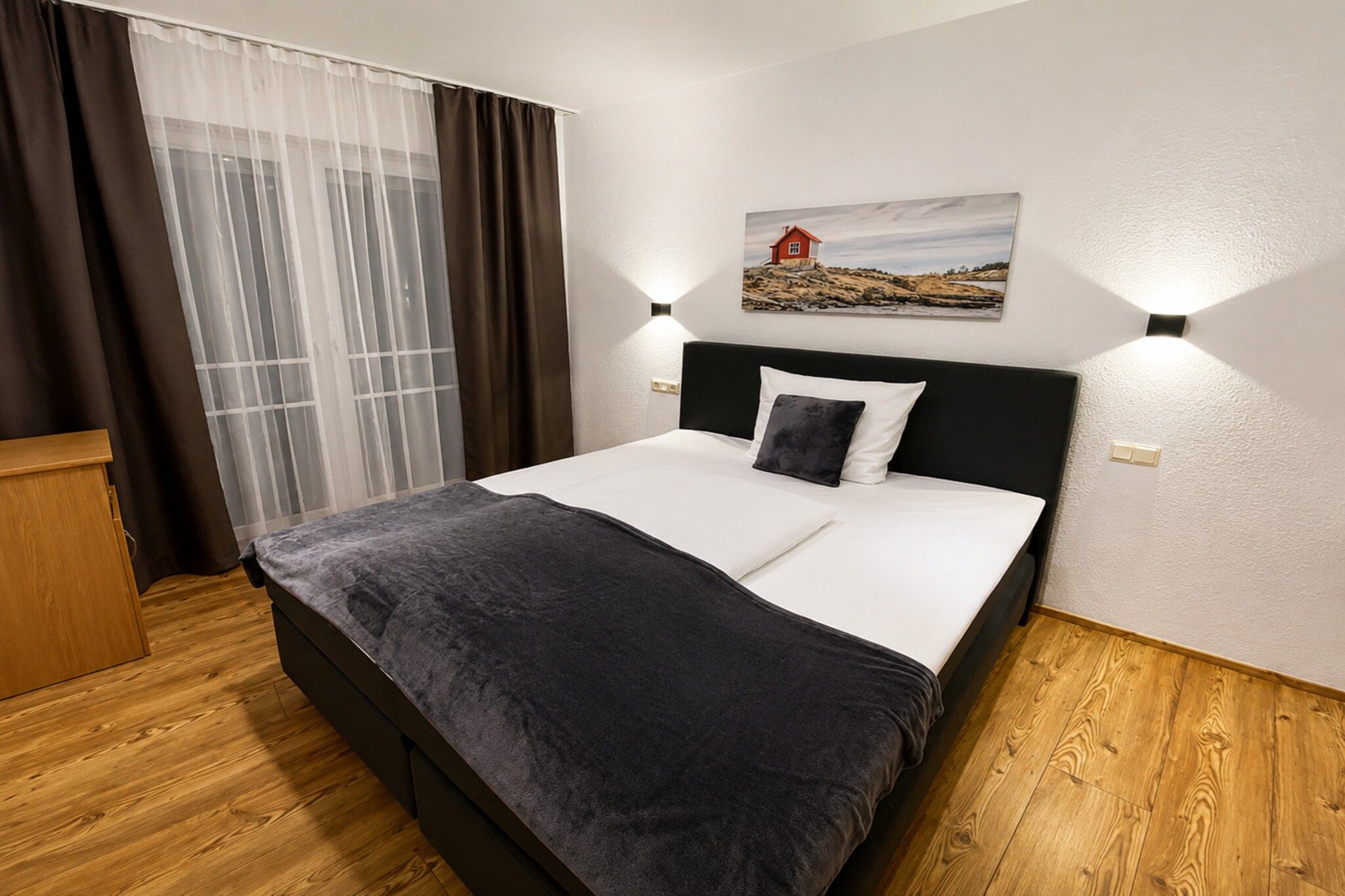A hotel room with a bed, a wooden floor, a window with curtains, and a painting on the wall.
