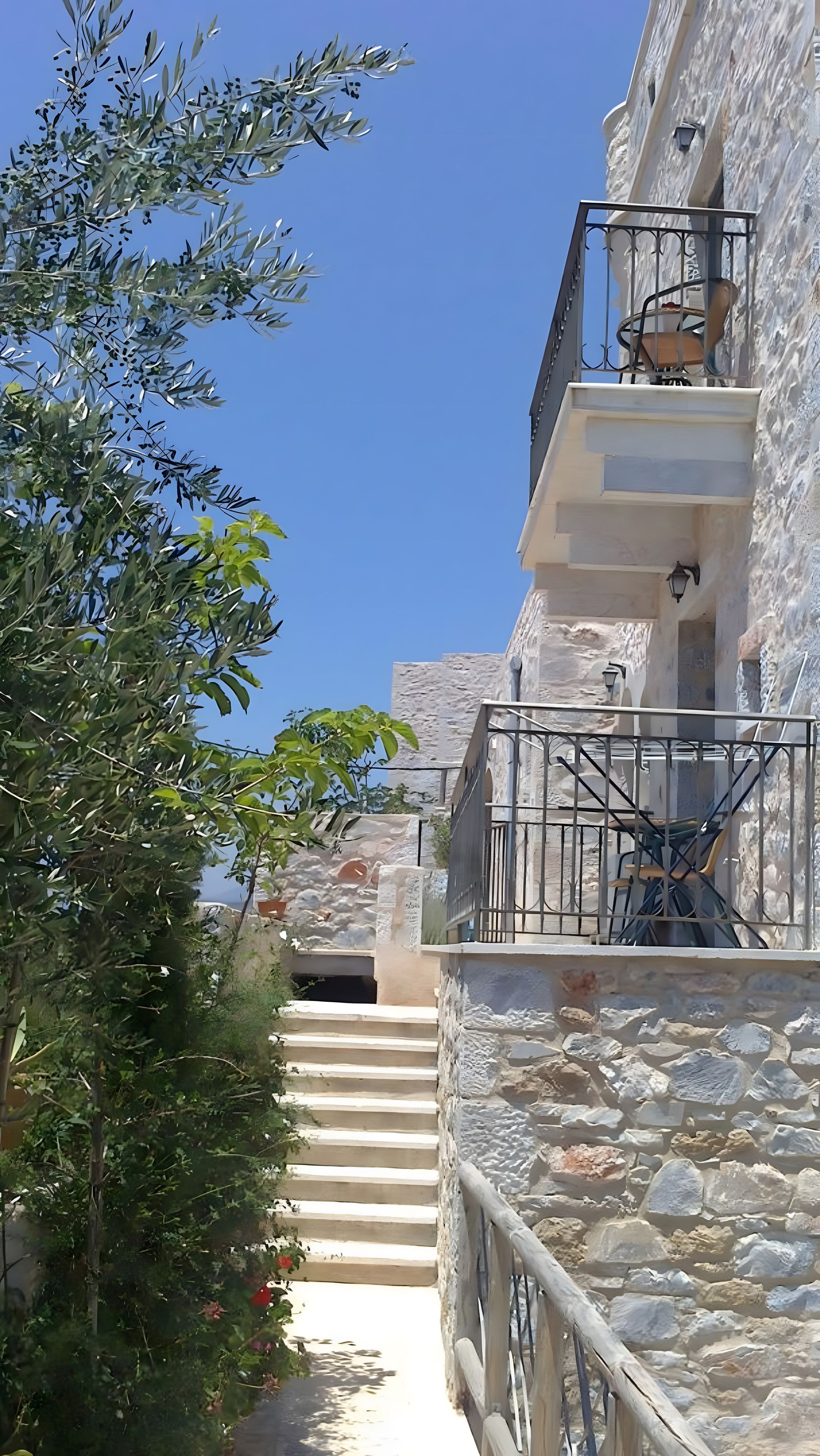 Ein Steinhaus mit einem Balkon und einer Treppe, die zum Eingang führt, vor einem wolkenlosen, blauen Himmel.