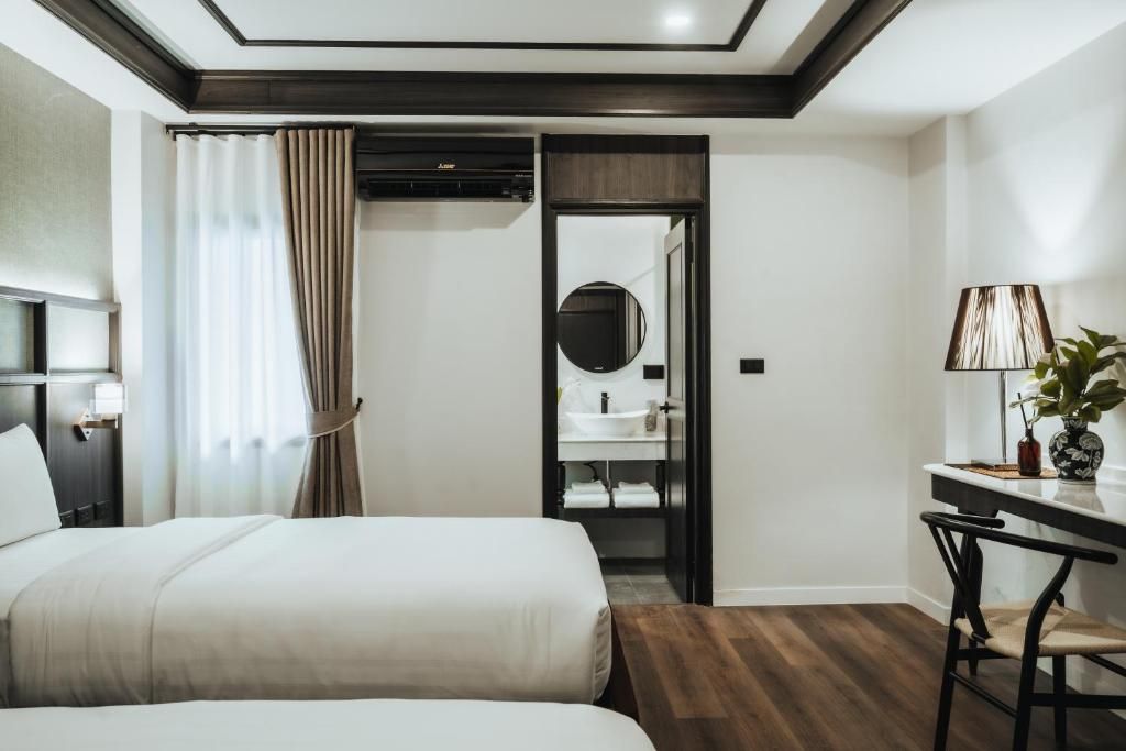 Habitación de hotel con dos camas, un escritorio, una silla, un espejo y un baño con lavamanos y toallero.