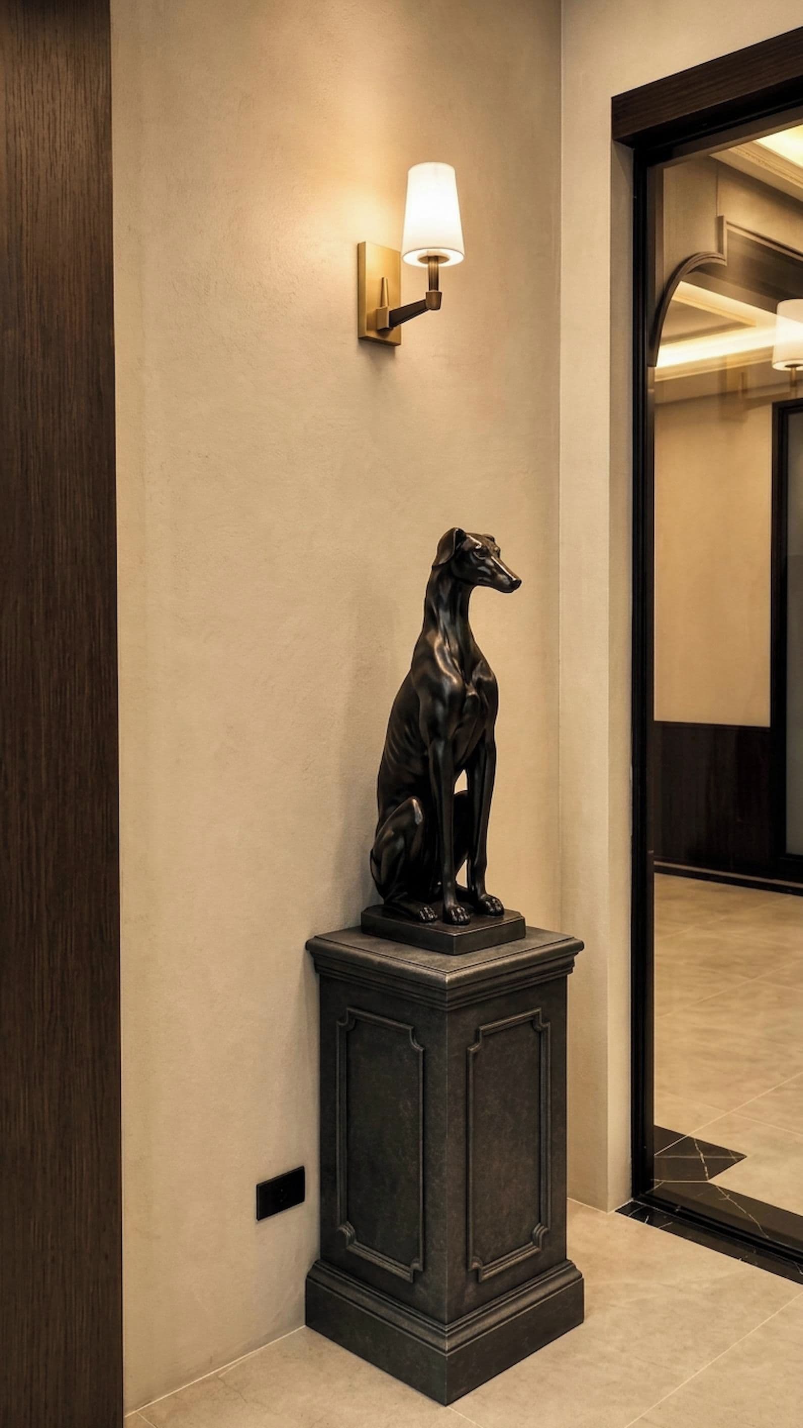 A black dog statue on a pedestal in a hallway with a wall-mounted light fixture.