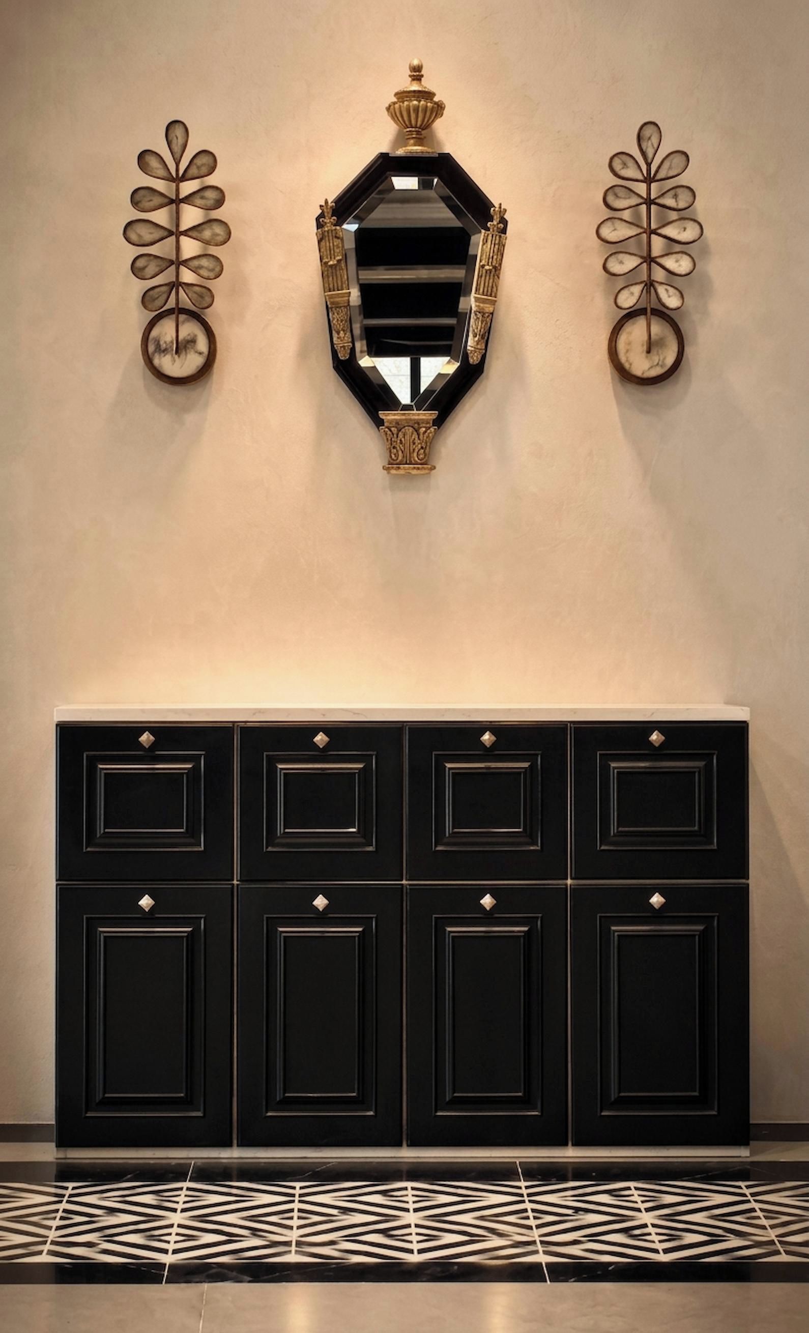 Un grand miroir mural, doté d’un cadre orné, est encadré de deux appliques murales aux motifs évoquant des feuilles. En dessous, une console noire, munie de nombreux tiroirs et de boutons décoratifs, est placée devant un sol à motifs.