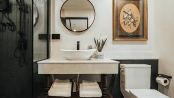 Une salle de bain avec un lavabo blanc, un miroir rond, une œuvre d’art encadrée et des toilettes.