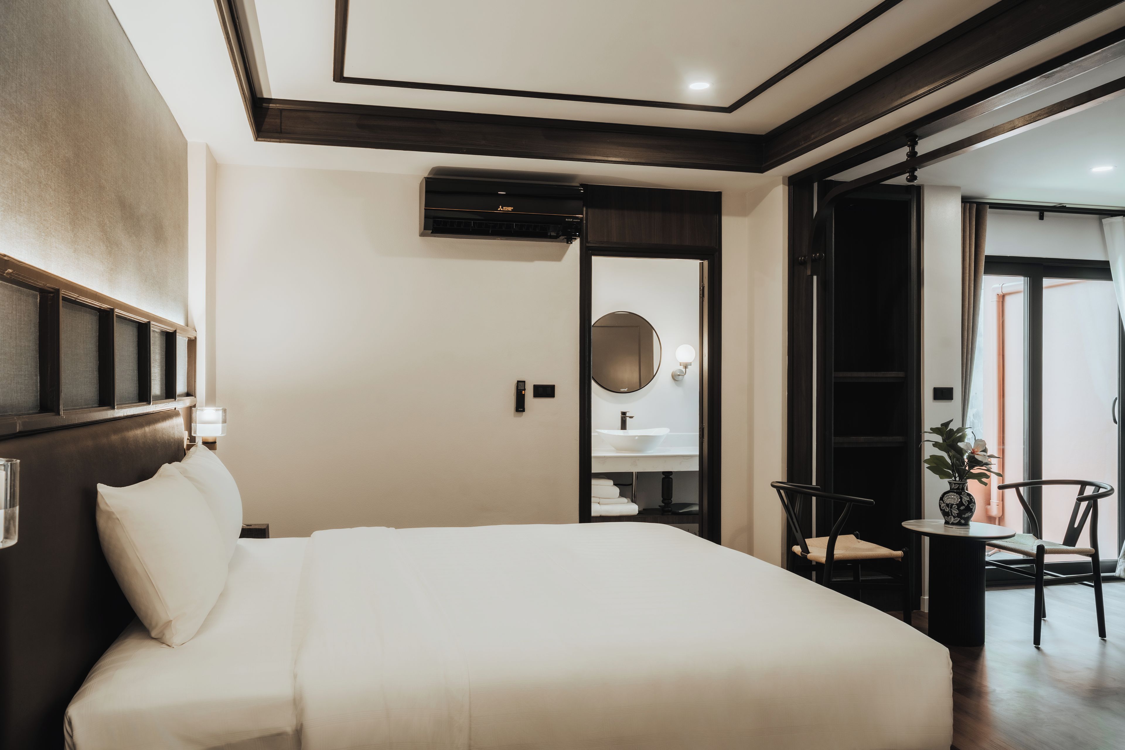 A hotel room with a large bed, a bedside table, a mirror, a small table with a chair, and a view of the outside.