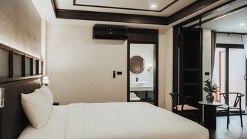 A hotel room with a large bed, a bedside table, a mirror, a small table with a chair, and a view of the outside.