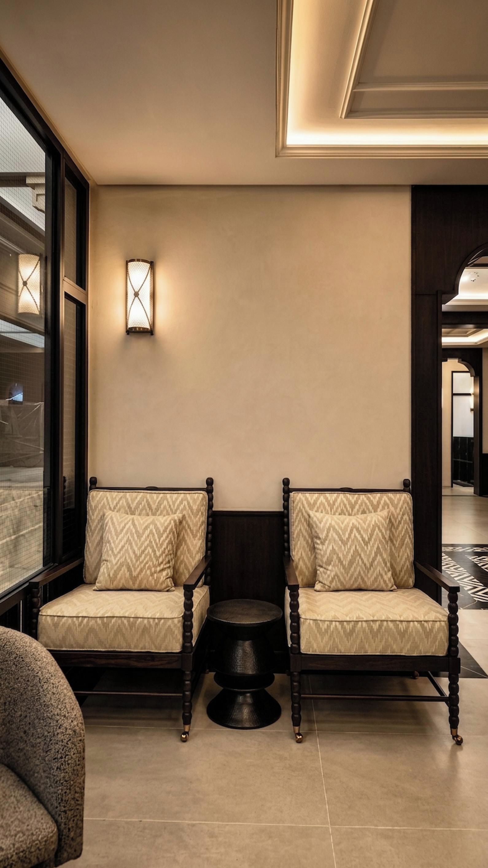 A modern hotel lobby with two upholstered chairs, a small round table, and a wall-mounted light fixture.