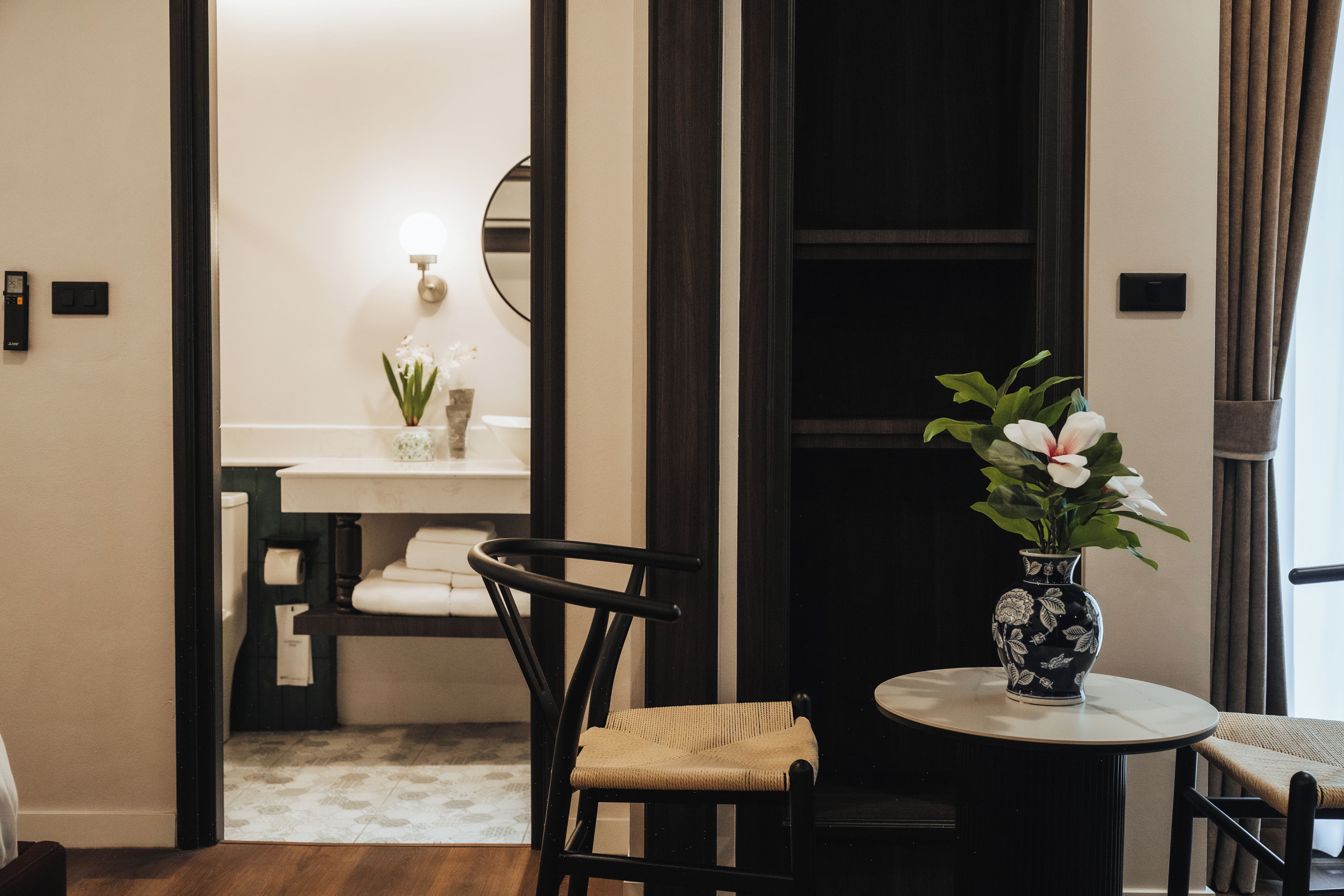 Habitación de hotel con una mesa redonda, una silla, un jarrón pequeño con flores, un espejo y una toalla en la pared.