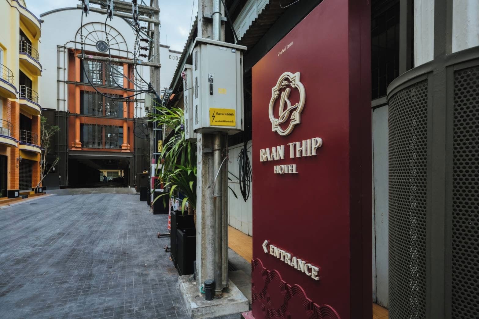 Vista de la calle de la entrada de un hotel con fachada color burdeos, un cartel con el nombre 'BAAN THIP HOTEL' y un camino despejado que lleva a la entrada.