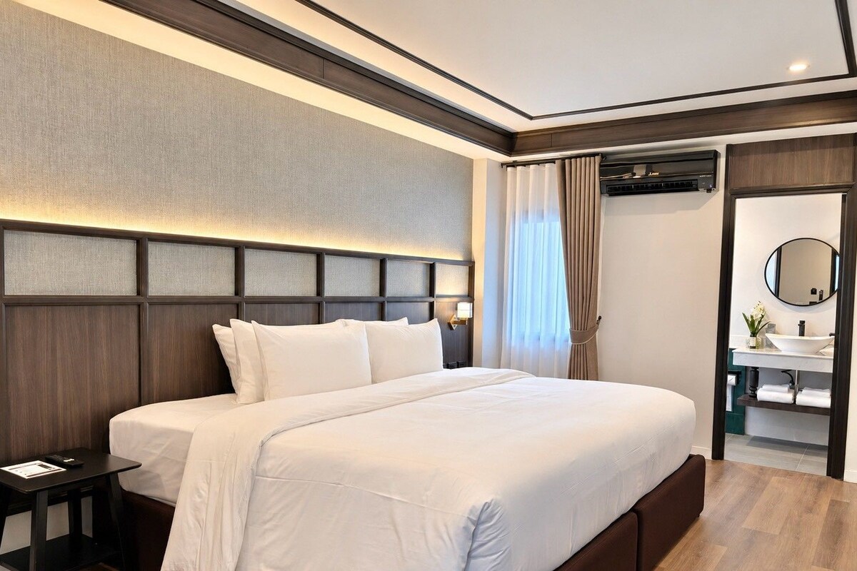 A hotel room with a large bed, a bedside table, a nightstand, a bathroom with a sink and mirror, and a window with curtains.