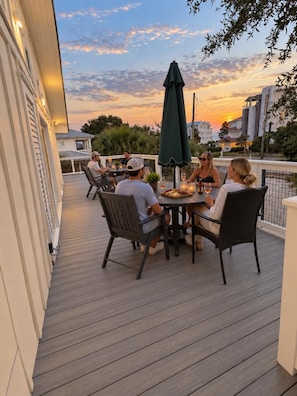 Eine Gruppe von Personen sitzt an einem Tisch auf einer Holzterrasse, im Hintergrund ein Sonnenuntergang.