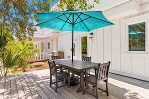 A wooden deck with a table and chairs, a blue umbrella, and a white building with a window and a fence. - Welcome to Salty Palm 🌴<br>Escape to Salty Palm, a spacious 6-bedroom 30A beach retreatjust steps from the beautiful white-sand beaches and emerald waters of the Emerald Coast! (Inlet Beach)