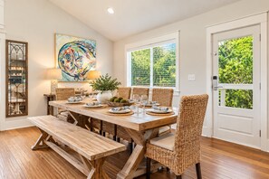A dining room with a wooden table set for four, surrounded by wicker chairs, a large painting, and a window with a view of greenery. - Welcome to Salty Palm 🌴<br>Escape to Salty Palm, a spacious 6-bedroom 30A beach retreatjust steps from the beautiful white-sand beaches and emerald waters of the Emerald Coast! (Inlet Beach)