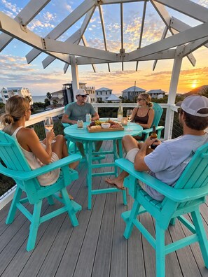 Eine Gruppe von Personen sitzt an einem runden Tisch auf einer Holzterrasse mit Blick auf das Meer und Häuser im Hintergrund.