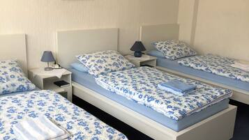 A hotel room with three single beds, each with a blue floral bedspread and matching pillows. There is a small bedside table with a lamp on each bed.
