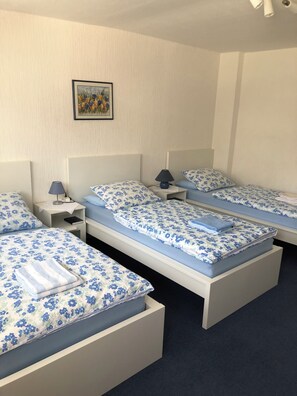A hotel room with three single beds, each with a blue floral bedspread and matching pillows. There is a small bedside table with a lamp on each bed.