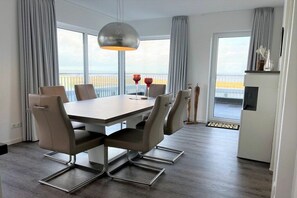 A modern dining area with a rectangular table, six chairs, a pendant light, and a view of the sea through large windows. - Holiday apartment with 1 bedroom (Wangerland)