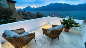 Eine Terrasse mit Korbmöbeln und einem Blumentopf, mit Blick auf einen See und Berge.
