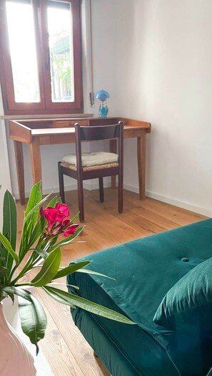 Ein Zimmer mit einem Holztisch, einem Stuhl, einem türkisfarbenen Sofa und einem Blumentopf mit grünen Blättern und roten Blüten.