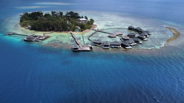 Una vista aerea di un'isola con bungalow sull'acqua collegati da passerelle, circondata da acque turchesi cristalline.