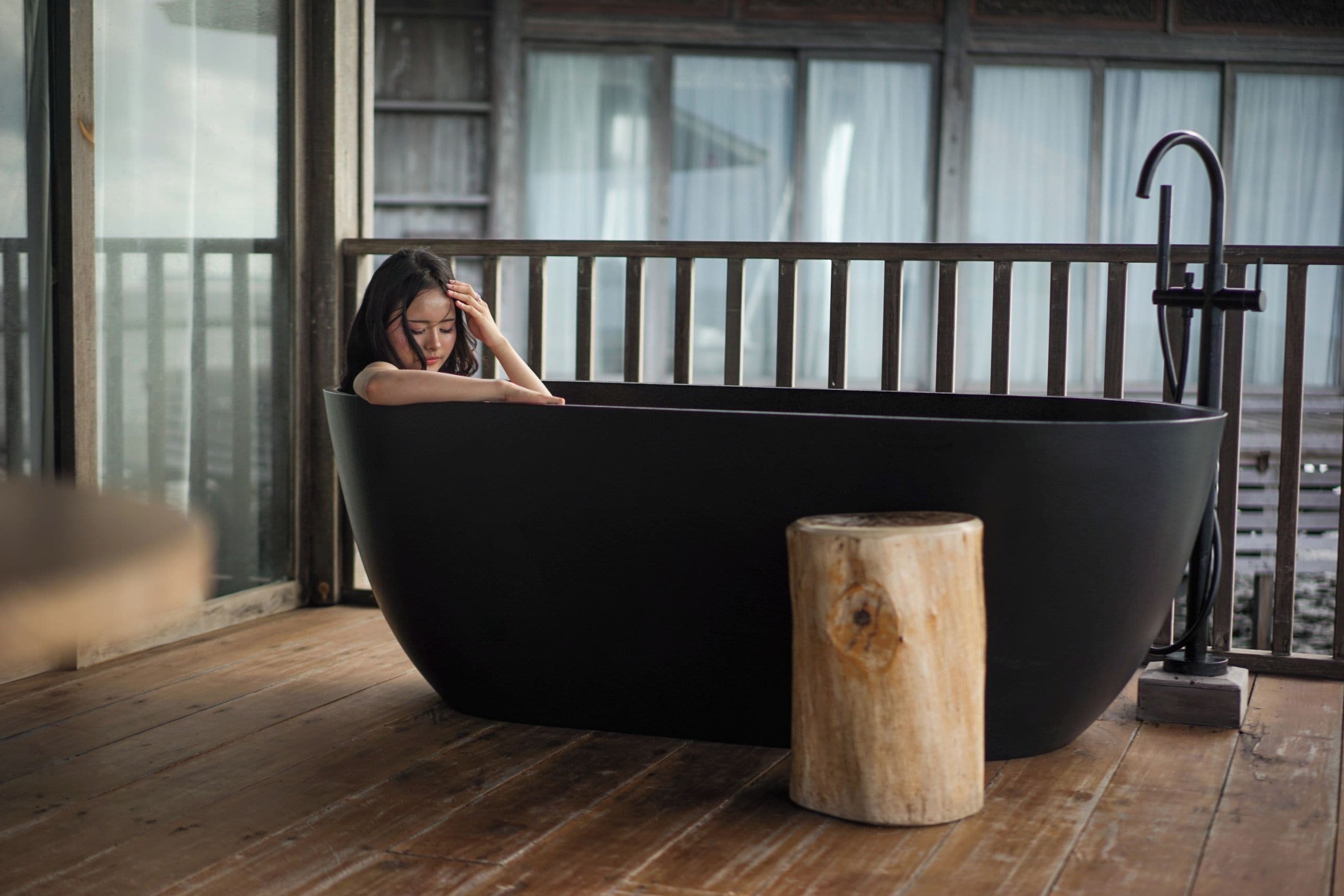 A person relaxing in a large black bathtub with a wooden stool beside it, on a wooden deck with a view of buildings through large windows.