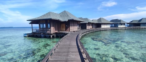 A wooden overwater bungalow with a covered walkway leading to it, set in clear turquoise waters.